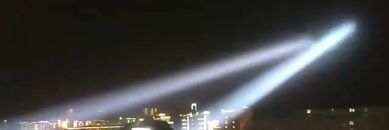 Two searchlight beams crossing in an X over a lit city skyline