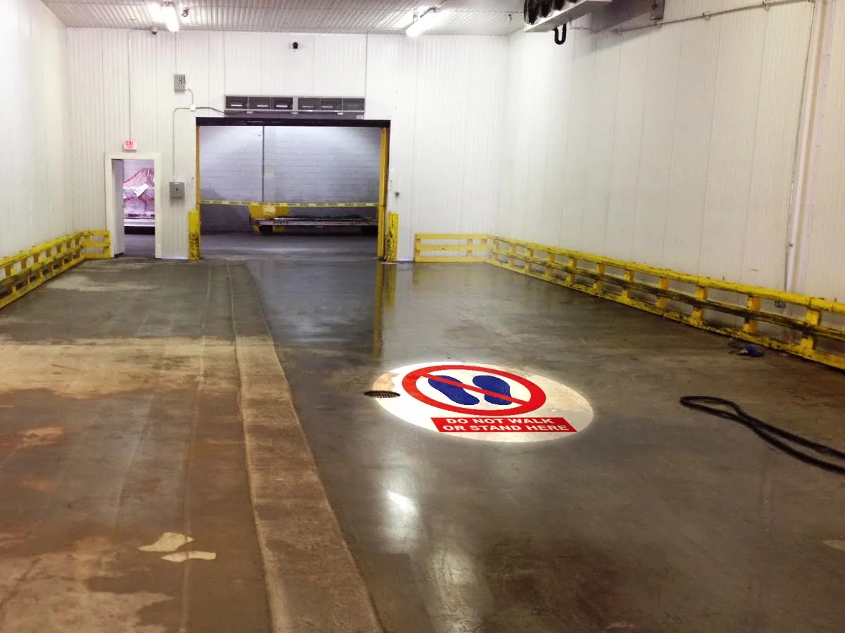DO NOT WALK OR STAND HERE safety sign projected on a cold-storage dock floor beside yellow guard rails